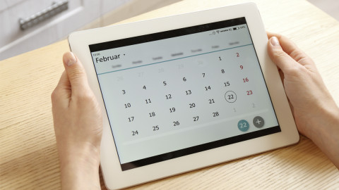 Zwei Hände halten ein Tablet, das eine Kalender-App anzeigt. 
