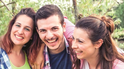 Zwei junge Frauen und ein junger Mann, untergehakt, schauen lachend in die Kamera.